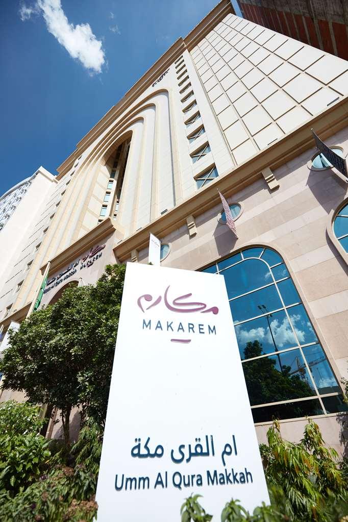 فندق مكارم أم القرى مكة المظهر الخارجي الصورة The photo shows the exterior of a building featuring a prominent sign that reads "MAKAREM." The design of the building is modern, with large windows and a mix of light-colored stone and other materials. There are decorative elements and flags on the 