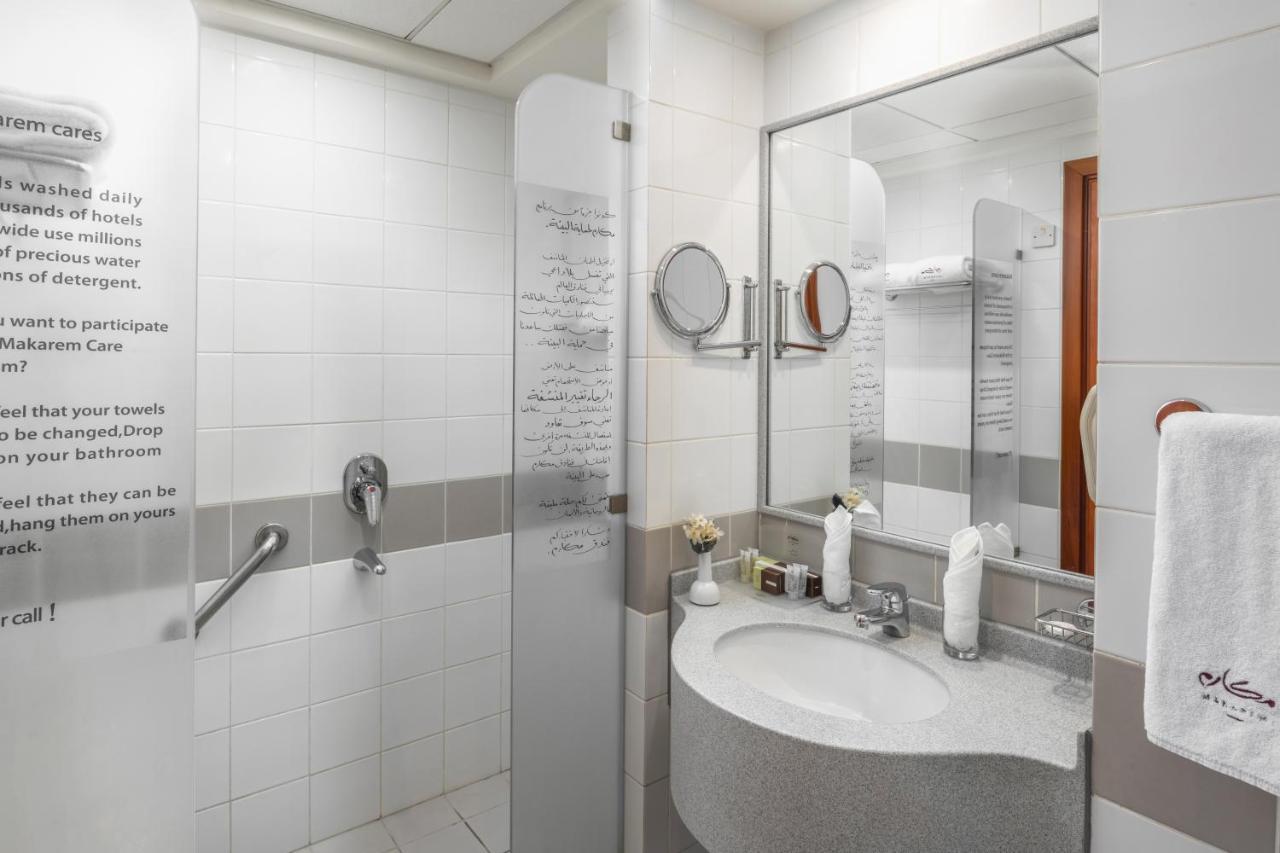 فندق مكارم أم القرى مكة المظهر الخارجي الصورة The photo shows a modern bathroom with a clean, minimalist design. It features a shower area separated by a glass panel, and there is a round mirror above a curved sink. The sink is accompanied by various toiletries and a hand towel. White tiles domi
