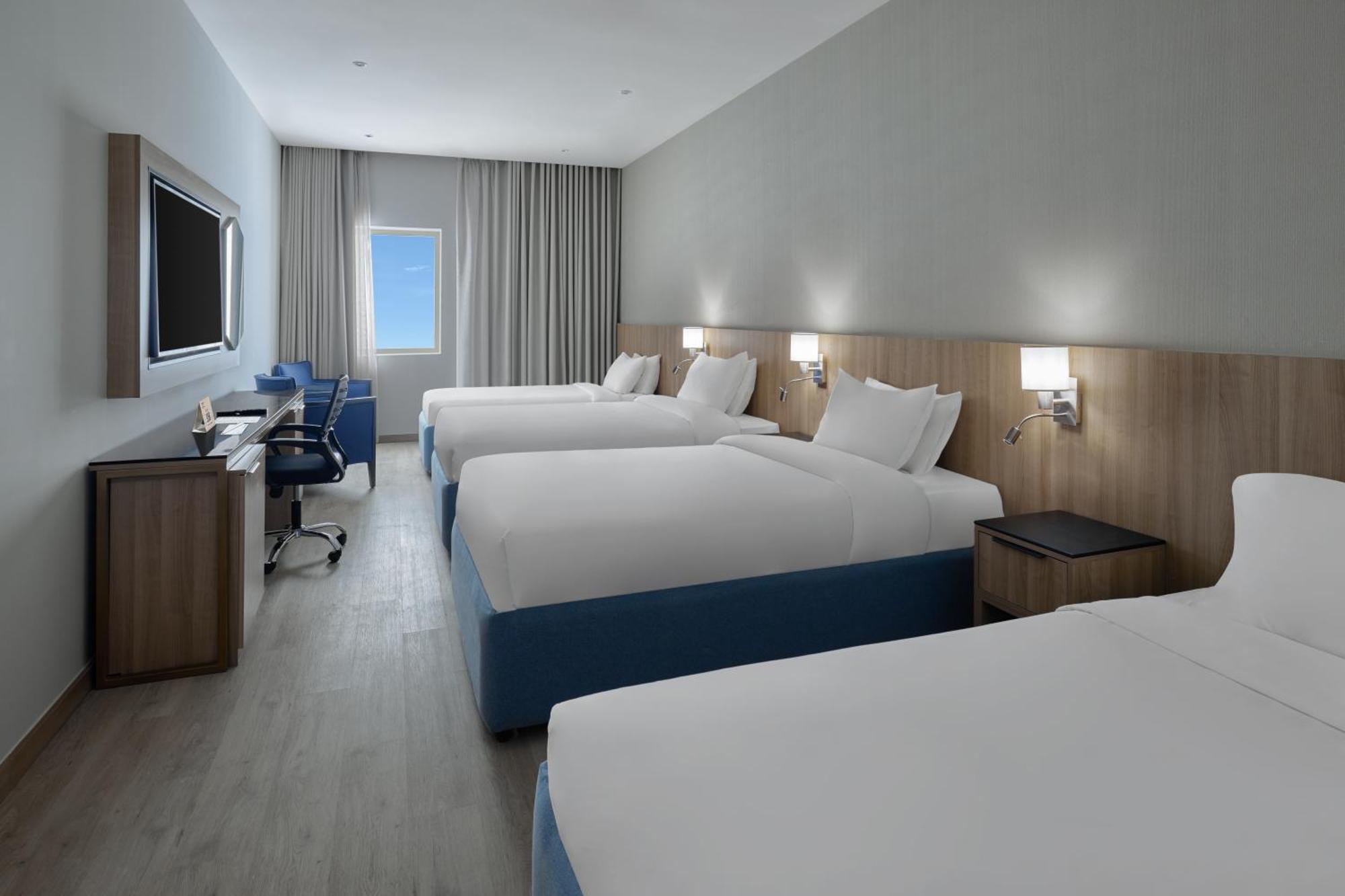 فندق مكارم أم القرى مكة المظهر الخارجي الصورة The photo shows a modern hotel room featuring four beds set up in a spacious layout. Each bed has white linens and blue accents. There is a large flat-screen TV mounted on the wall, and a desk with a chair is positioned next to it, providing a worksp