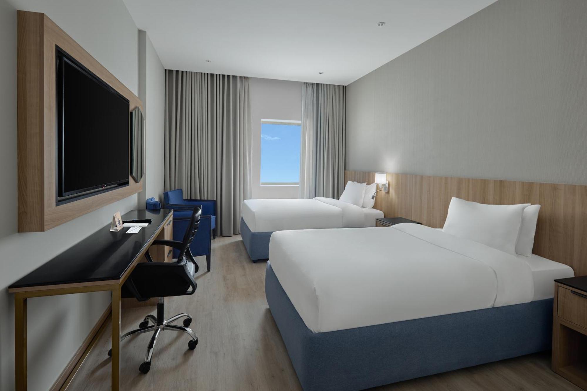 فندق مكارم أم القرى مكة المظهر الخارجي الصورة The photo shows a hotel room featuring two beds with white linens and blue bases. There is a modern desk and chair positioned against one wall, with a large flat-screen television mounted above it. Natural light comes in through a window covered by s