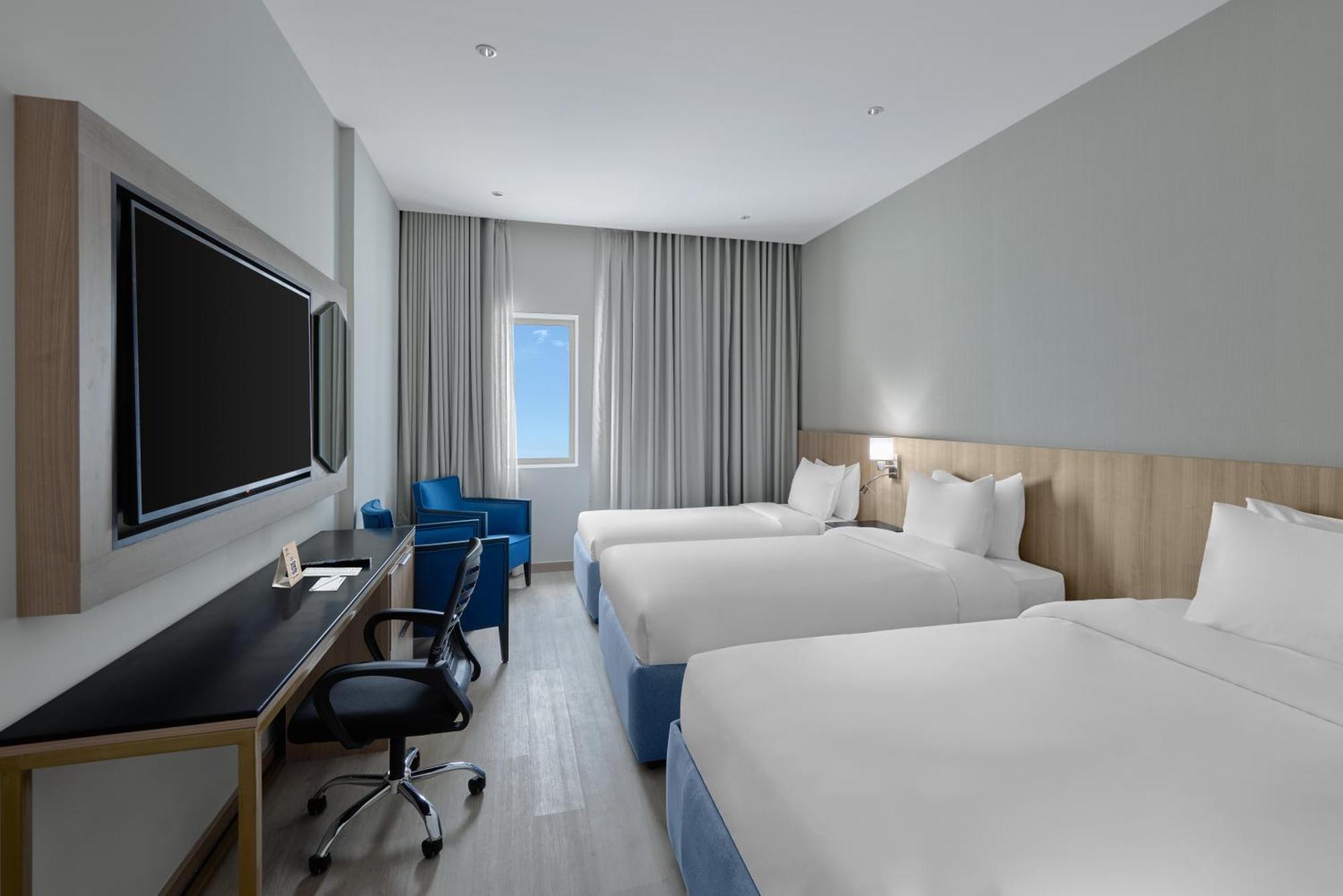 فندق مكارم أم القرى مكة المظهر الخارجي الصورة The photo shows a modern hotel room featuring two twin beds with white bedding. There's a light wood paneling behind the beds, and a large flat-screen TV mounted on the wall across from the beds. A comfortable blue armchair and a small desk with an e