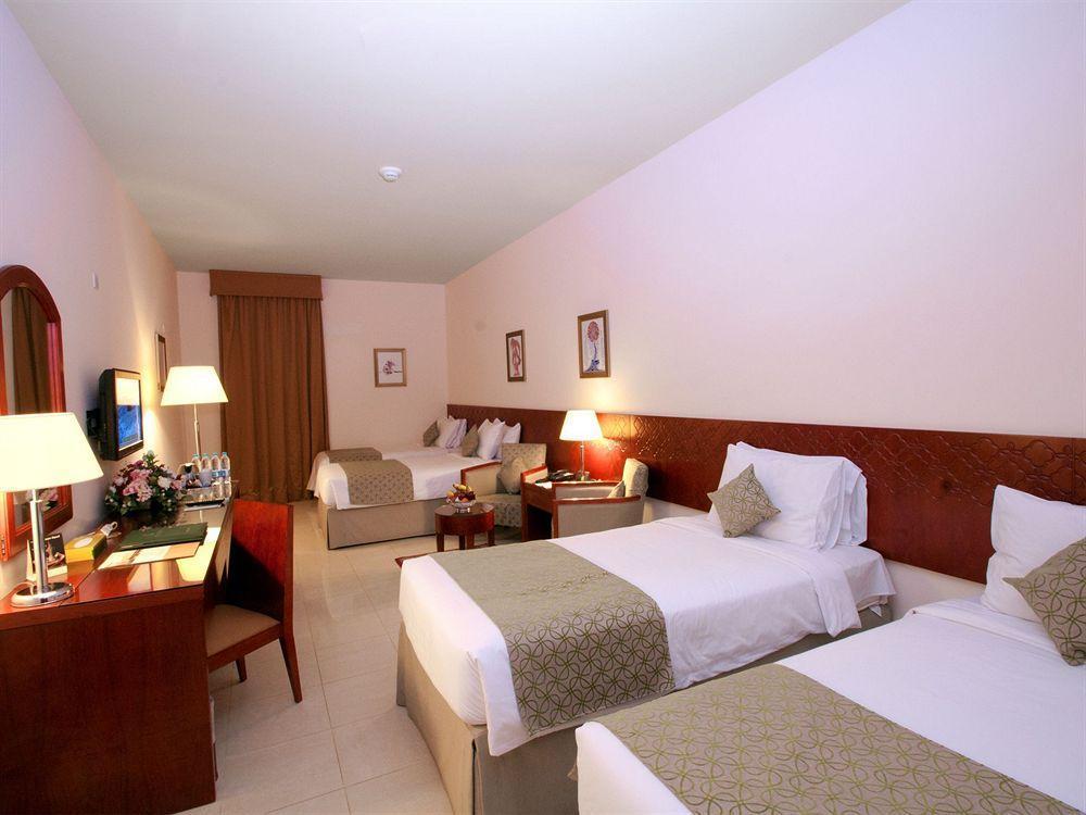 فندق مكارم أم القرى مكة المظهر الخارجي الصورة The photo shows a hotel room with a light, neutral color scheme. There are two twin beds on one side, each with white linens and decorative throw pillows. A bedside table with a lamp is positioned between the beds. On the other side of the room, ther