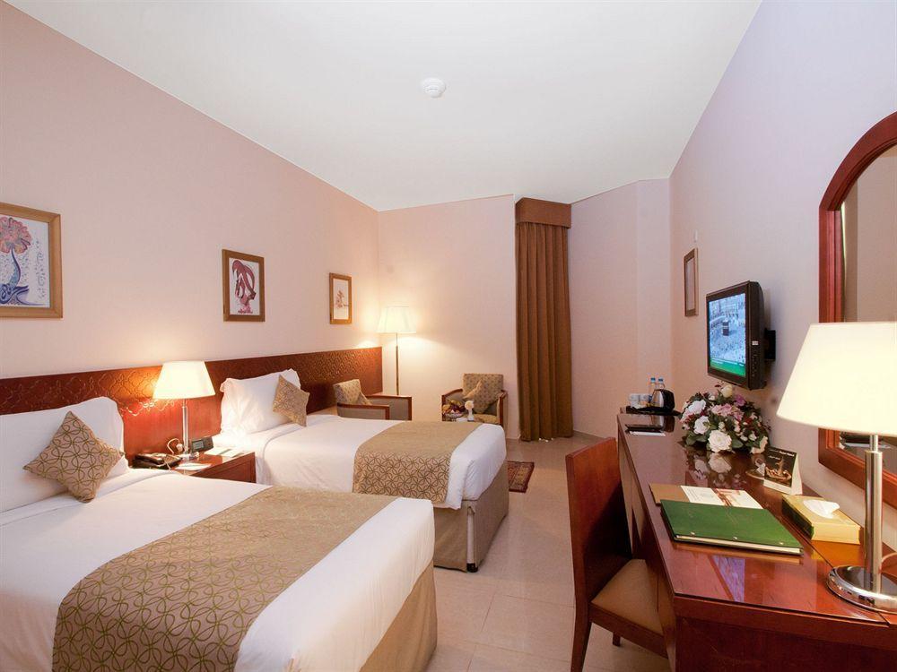 فندق مكارم أم القرى مكة المظهر الخارجي الصورة The photo shows a hotel room featuring two single beds with light-colored bedding and decorative throw pillows. There are bedside tables with lamps next to each bed, and wall art is displayed above the beds. A chair is positioned near the window, and