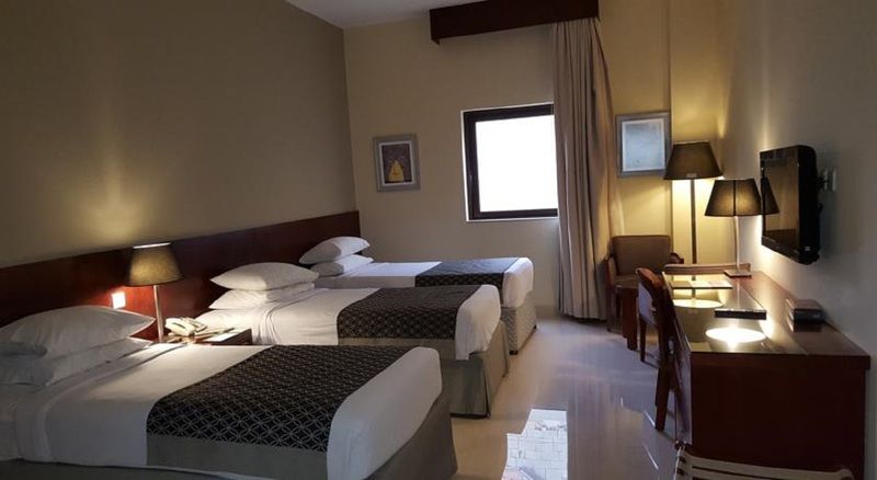 فندق مكارم أم القرى مكة المظهر الخارجي الصورة The photo shows a hotel room with two twin beds covered in white and patterned bedding. The walls are painted in a soft color, and there's a window with light-colored curtains. A small desk with a chair is positioned near the window, and there are tw