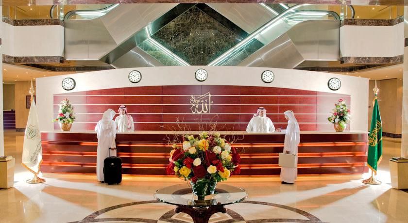 فندق مكارم أم القرى مكة المظهر الخارجي الصورة The photo depicts a luxurious hotel lobby. In the center, there is a reception desk made of wood with a decorative logo on it. Behind the desk, there are several clocks displaying different times, indicating an international setting. The reception ar