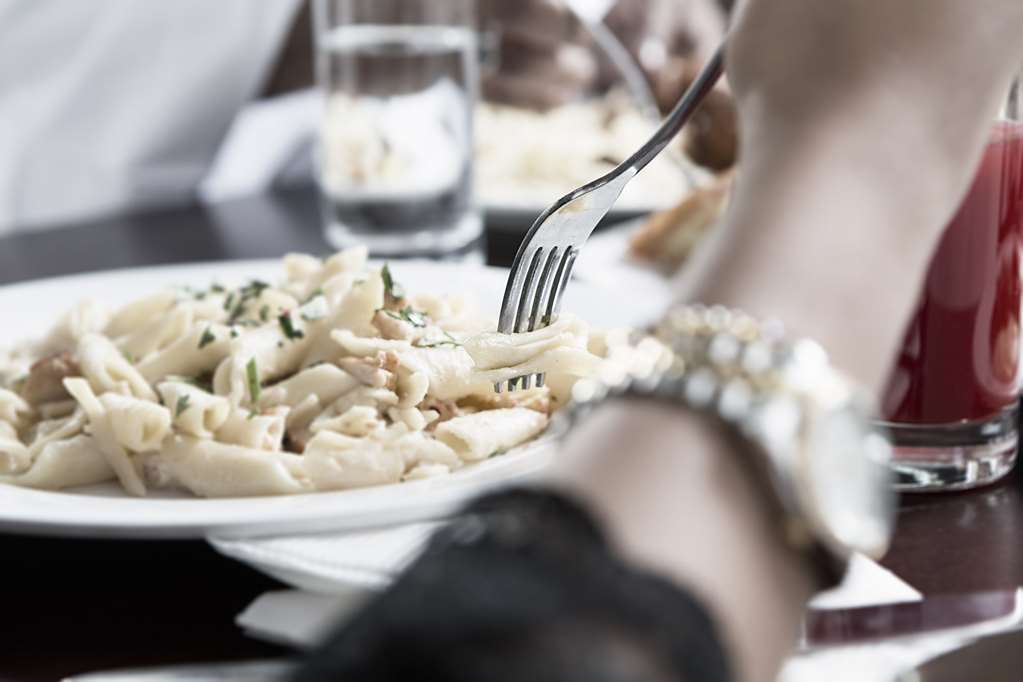 فندق مكارم أم القرى مكة المطعم الصورة The photo shows a close-up of a plate of pasta garnished with herbs, with a fork poised above it, suggesting someone is about to take a bite. To the side, there is a hand wearing a watch, and in the background, there are glasses of water on the table