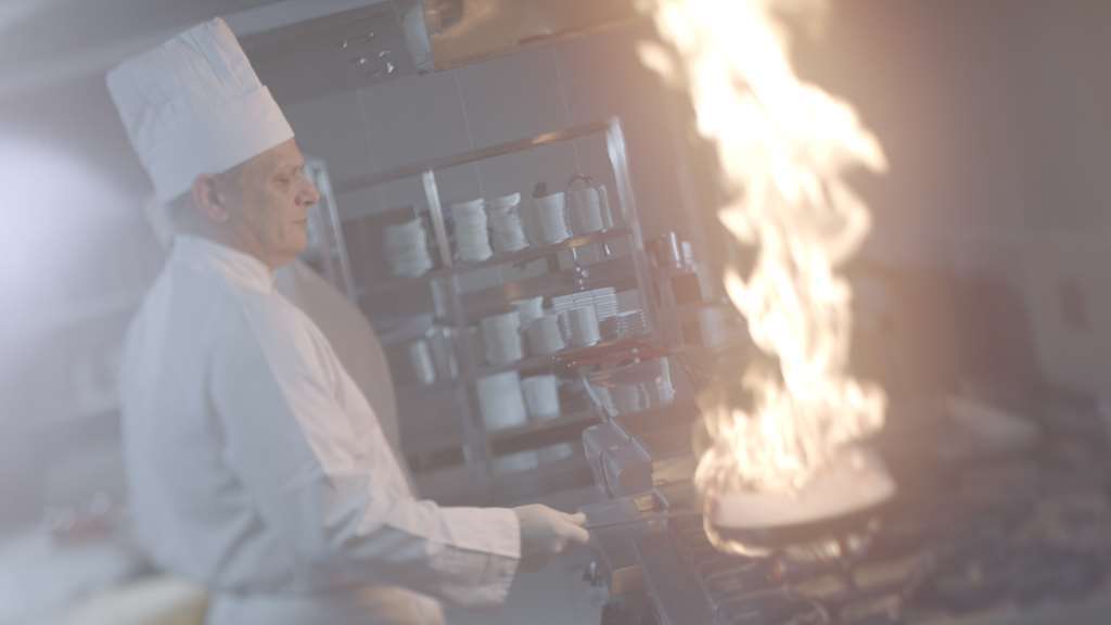 فندق مكارم أم القرى مكة المطعم الصورة The image shows a chef in a kitchen setting. He is wearing a white chef's uniform and a traditional chef's hat. There is a large flame erupting from a pan on the stove, which suggests that he is likely engaged in cooking that involves high heat or fl