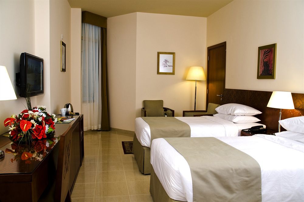 فندق مكارم أم القرى مكة المظهر الخارجي الصورة The photo shows a hotel room featuring two twin beds with white bedding and olive green accents. Next to the beds is a small bedside table with a lamp. There is a comfortable chair positioned nearby. A television is mounted on the wall, and there's a