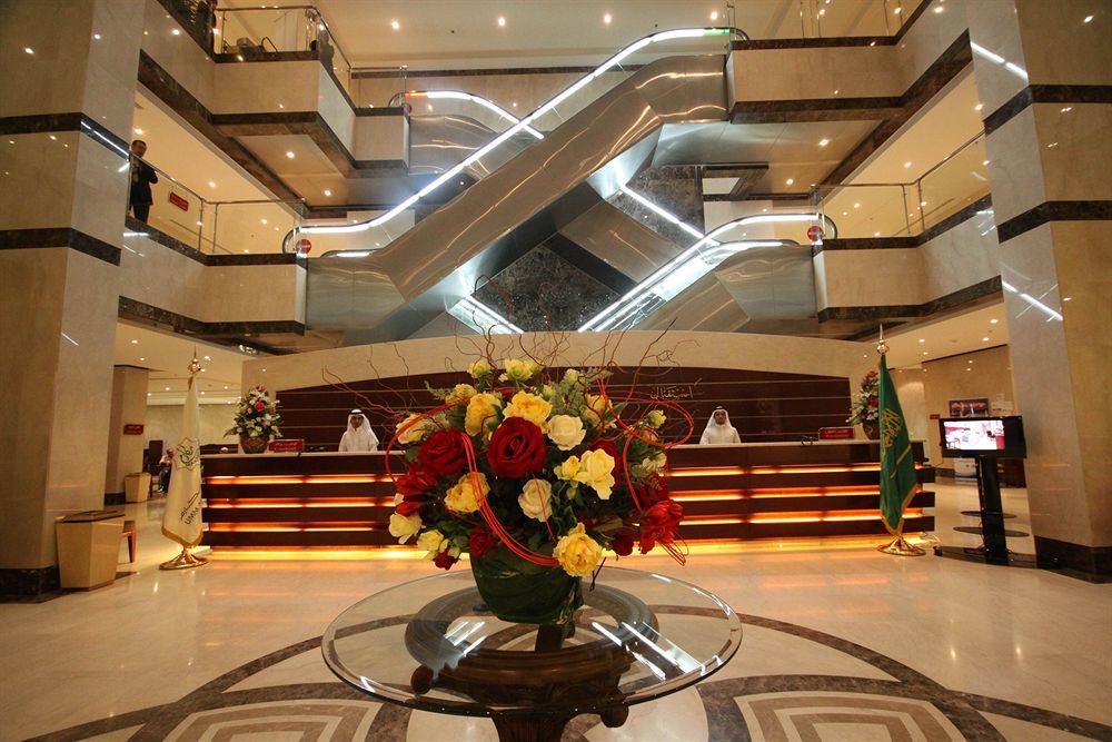 فندق مكارم أم القرى مكة المظهر الخارجي الصورة The photo shows a modern hotel lobby. In the foreground, there is an elegant floral arrangement featuring various colorful flowers in a decorative vase. In the background, a reception desk is visible, staffed by individuals in uniform. Two escalators