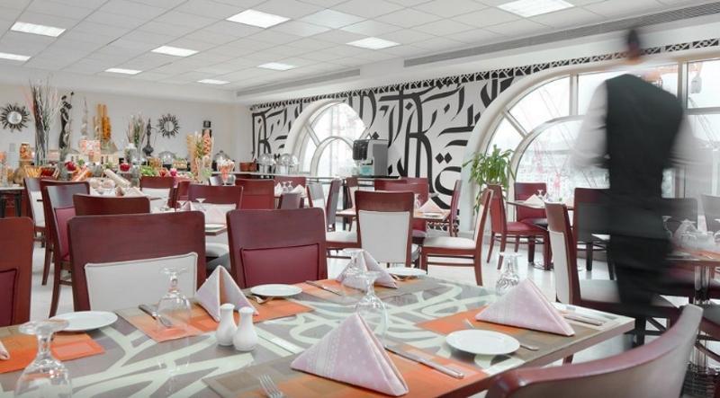 فندق مكارم أم القرى مكة المظهر الخارجي الصورة The photo shows the interior of a restaurant. It features a modern design with a combination of wooden and white furniture. The tables are set with white plates and folded napkins, arranged neatly. There are decorative touches such as small plants an